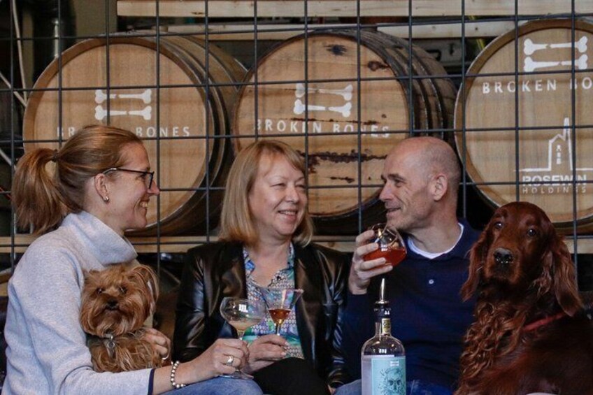 Three people holding drinks, a dog, and a bottle of liquor in front of wooden barrels with text. 1
