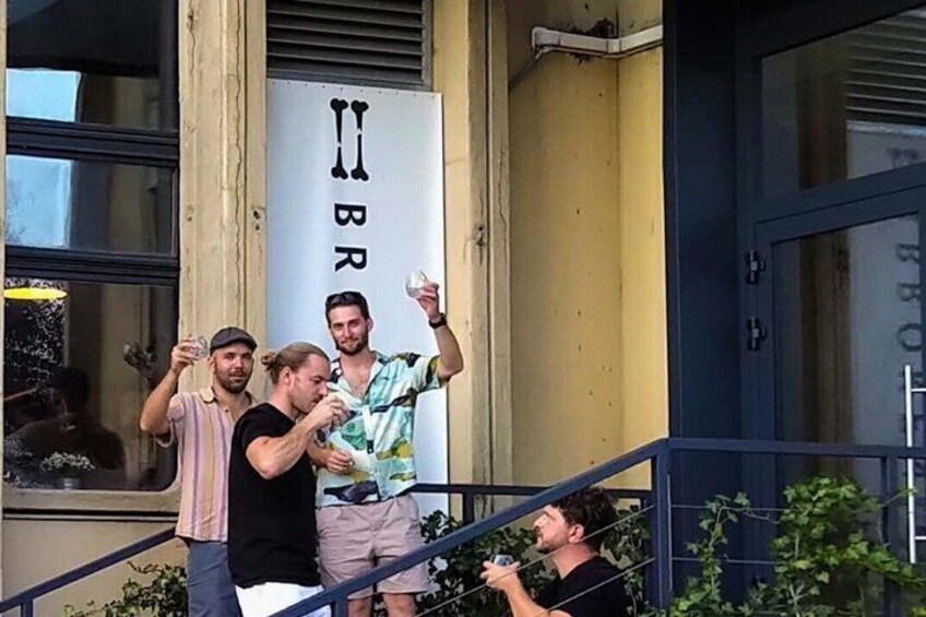A group of people on a balcony, some holding drinks, with a building featuring a sign with 'BR' visible. 5