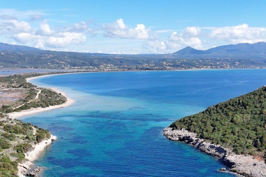 A coastal landscape with a clear blue sea, a sandy beach, and a rocky outcrop with vegetation. 1