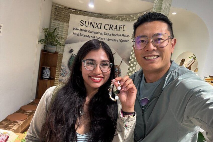 Two people in a craft store with a sign that reads 'SUNXI CRAFT Handmade Everything | Todos Hechos Mano | song Brocade Silk | Miao Embroidery | Quartz'. 3