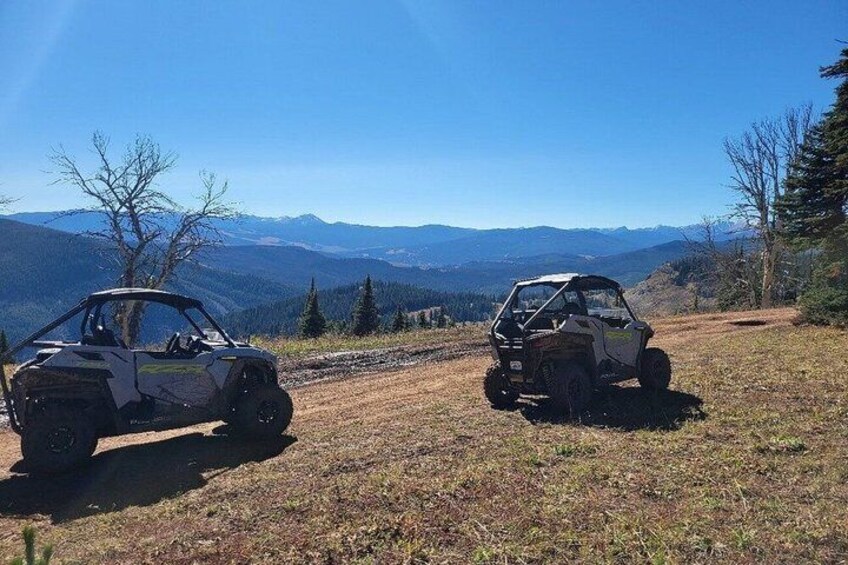Two off-road vehicles parked on a grassy hillside with a mountainous backdrop. 2