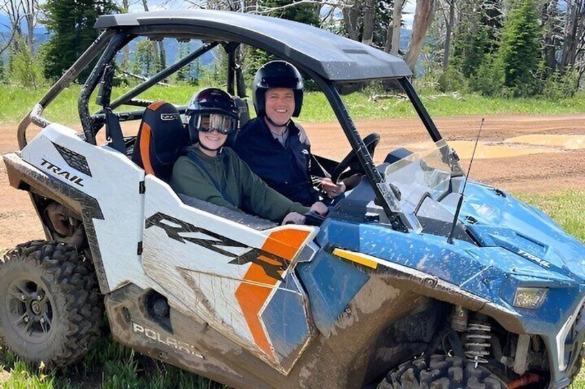 Two people in a Trail brand off-road vehicle on a dirt road. 1