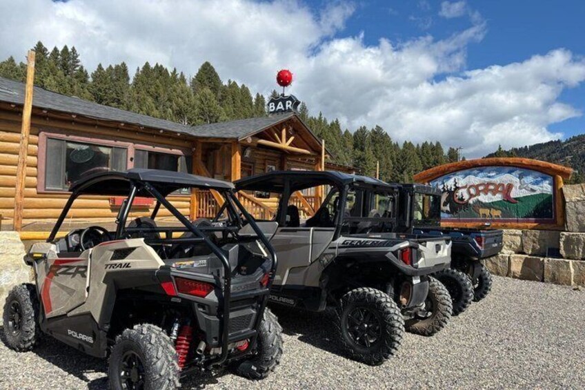Two Polaris RZR vehicles parked in front of a wooden cabin with a mountain mural. 3