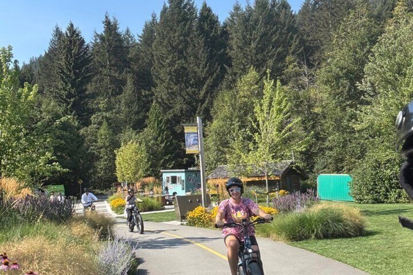 Cyclists on a paved path with a forested backdrop and a small cabin. 5