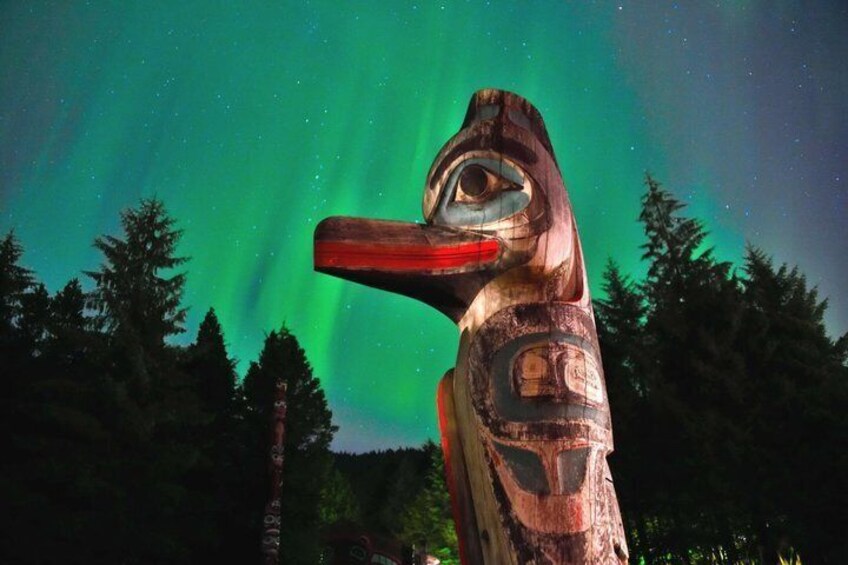 A totem pole with a carved face and a long beak, set against a forest background with the aurora borealis in the sky. 1