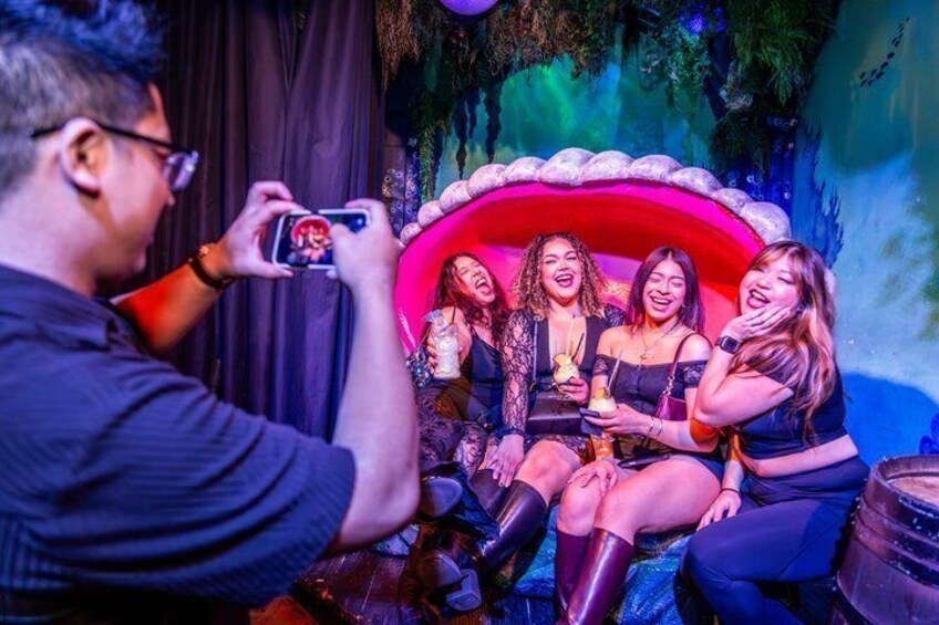 A group of people sitting on a couch with a red backdrop, a man taking a photo, and a bar area in the background. 1