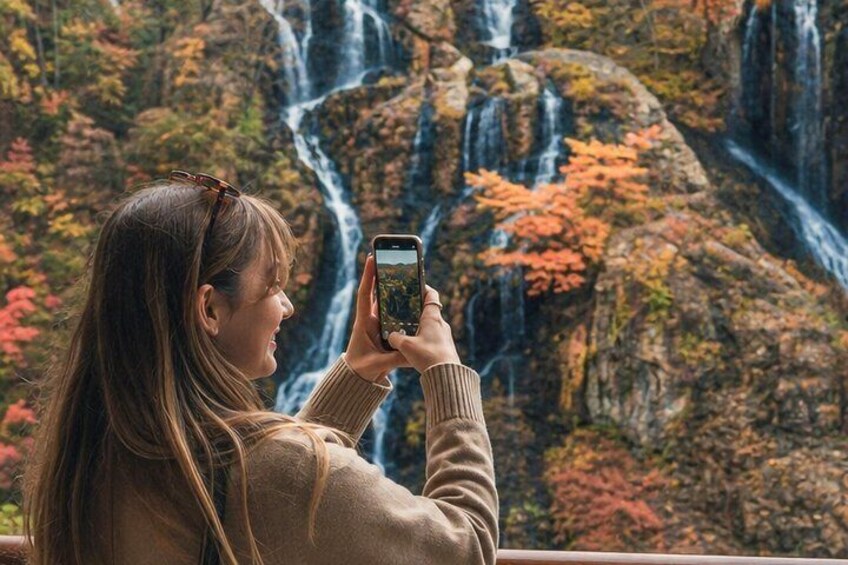 A person taking a photo of a waterfall with autumn foliage in the background. 5