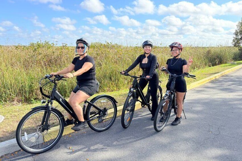 Three people on bicycles on a paved road with grass and wildflowers on the sides. 1