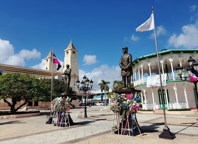 A plaza with a statue, a clock tower, and a building with arches. 5