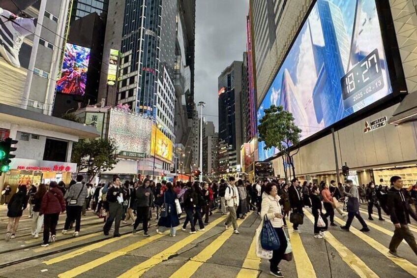 A busy urban street with pedestrians crossing a zebra crossing, tall buildings, and illuminated billboards. 1