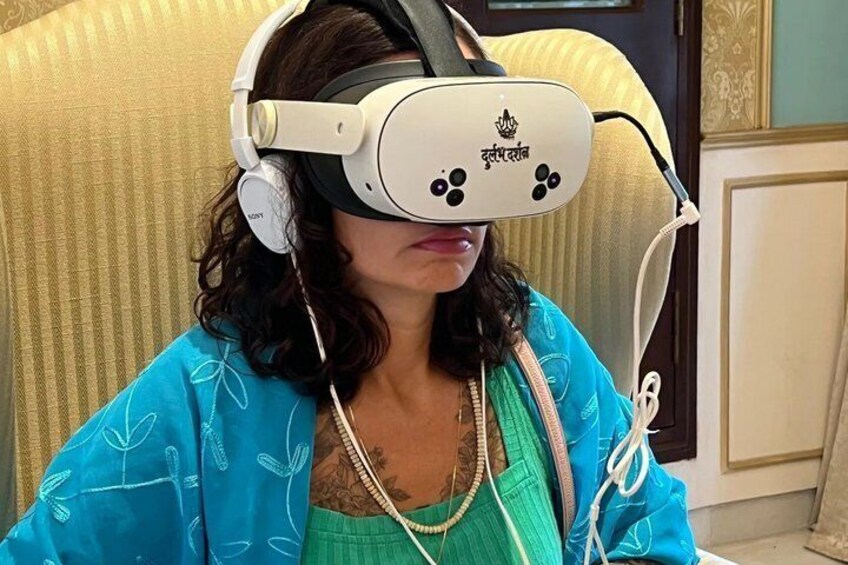 A person wearing a VR headset and headphones, seated in a room with patterned wallpaper and a decorative chair. 1