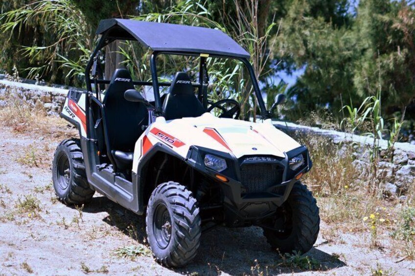 A four-wheeled off-road vehicle with a raised suspension, large tires, and a protective canopy, parked on a dirt path with vegetation in the background. 3