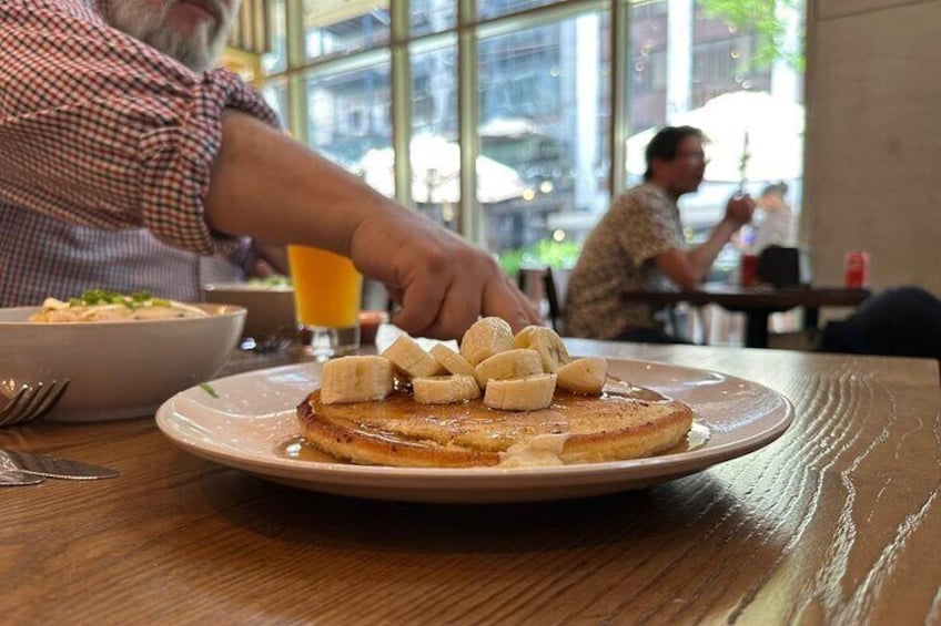 A breakfast plate with pancakes topped with bananas and syrup, a glass of orange juice, and a bowl of cereal on a wooden table in a cafe. 1