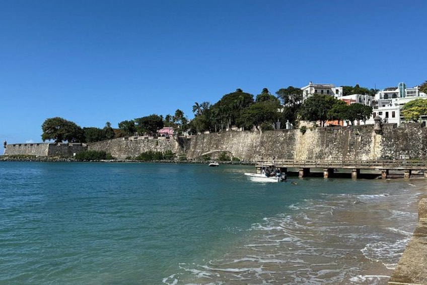 A coastal scene with a stone wall, a boat on the water, and buildings on a hill. 5