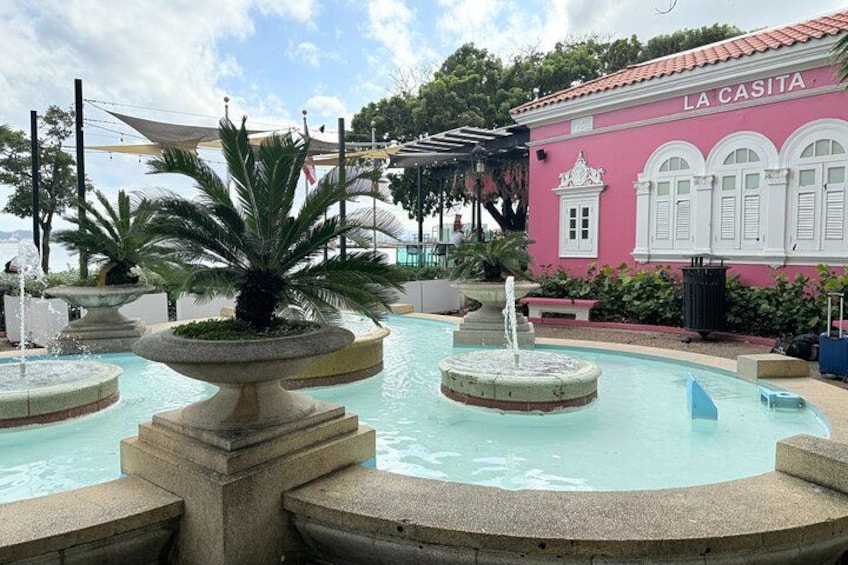 Una fuente con palmeras, un edificio rosa con la inscripción 'LA CASITA' y un cielo azul despejado. 4