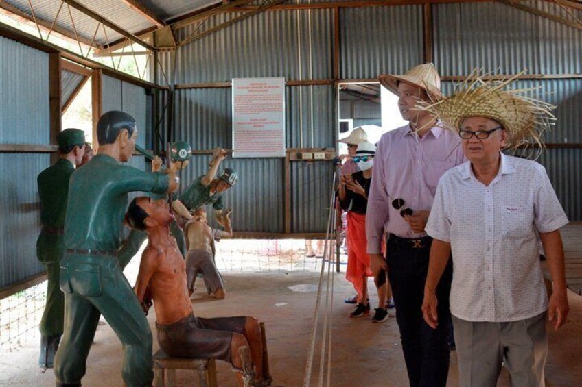 A group of people in a covered area with a corrugated metal roof, some wearing hats and others in casual clothing. 5