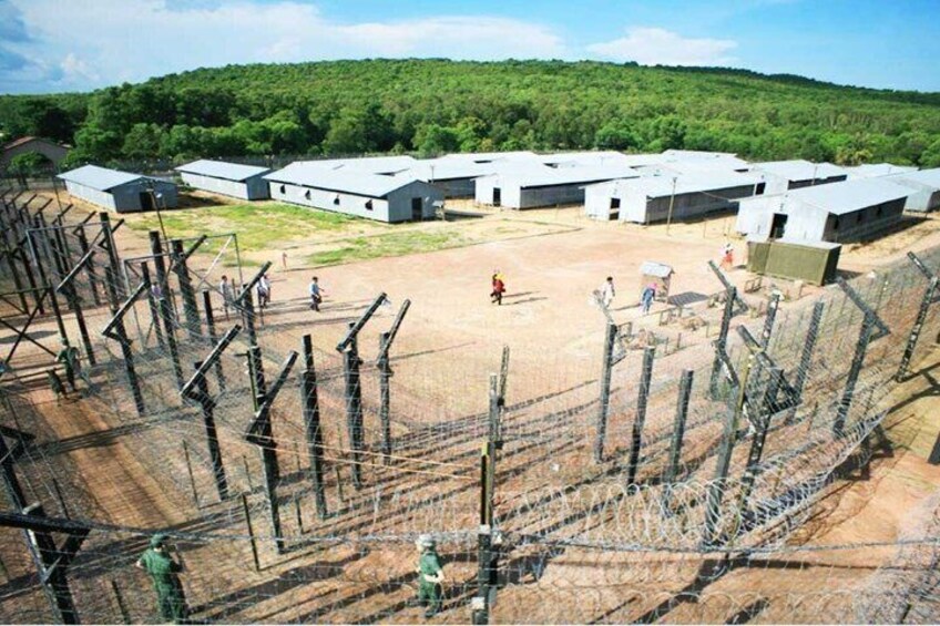 A fenced compound with buildings, a dirt area, and a forested hill in the background. 2