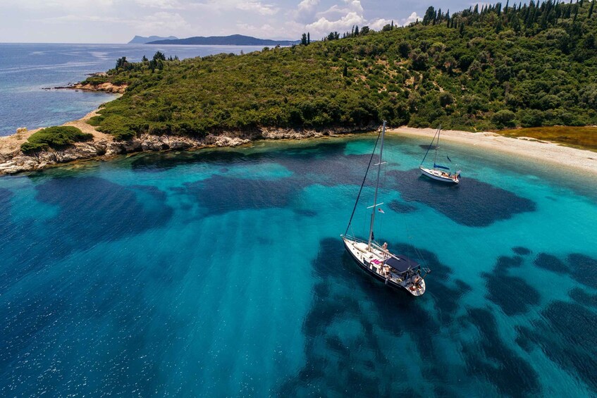A coastal landscape with clear turquoise waters, two sailboats anchored near the shore, and a forested hill in the background. 4