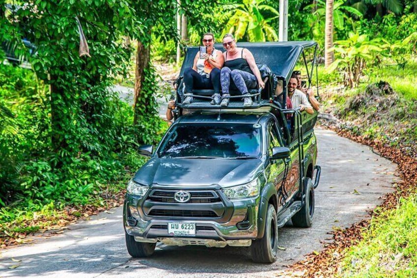 A Toyota pickup truck with a roof rack carrying four people on a paved road surrounded by lush greenery. 1