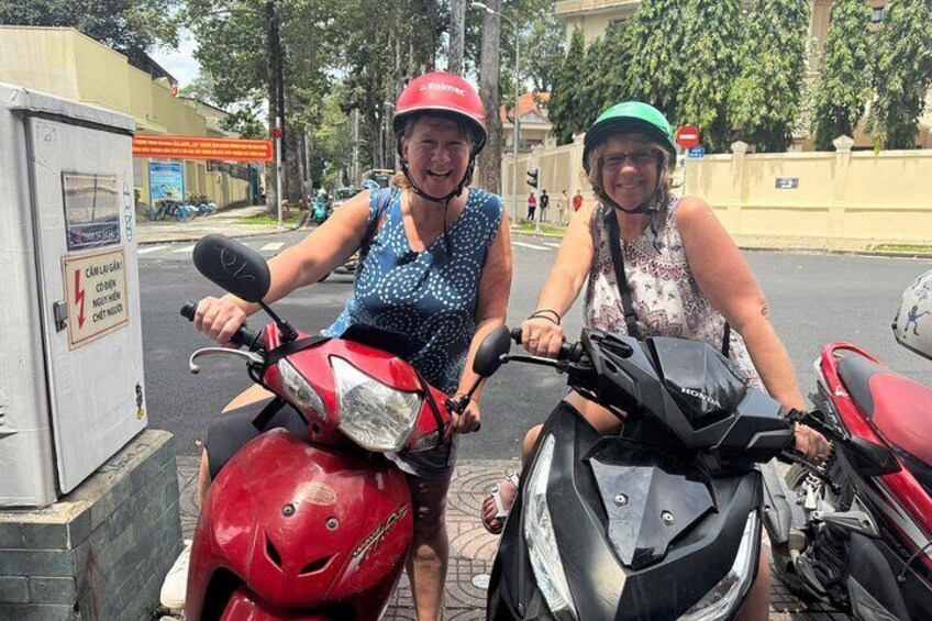 Two people on scooters, one red and one black, parked on a street with buildings and trees in the background. 1