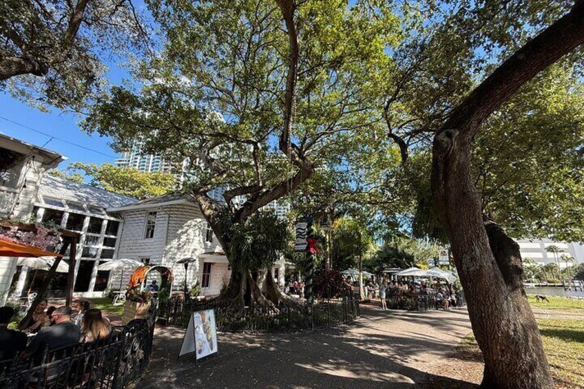 A tree-lined street with a white building, outdoor seating, and a market area. 4