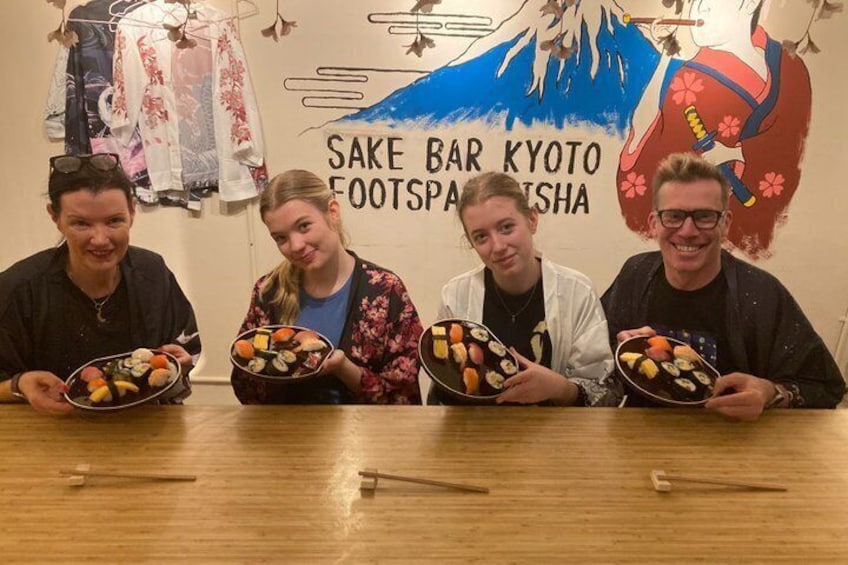 Four people holding plates of sushi at a table with a backdrop of a wall with Japanese text and a mountain illustration. 2
