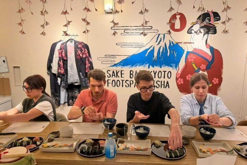 Four people seated at a table with plates of sushi, a wall with a Japanese flag and a mountain illustration, and a sign reading 'SAKE BAR KAWAKYOTO FOOTSPA'. 3