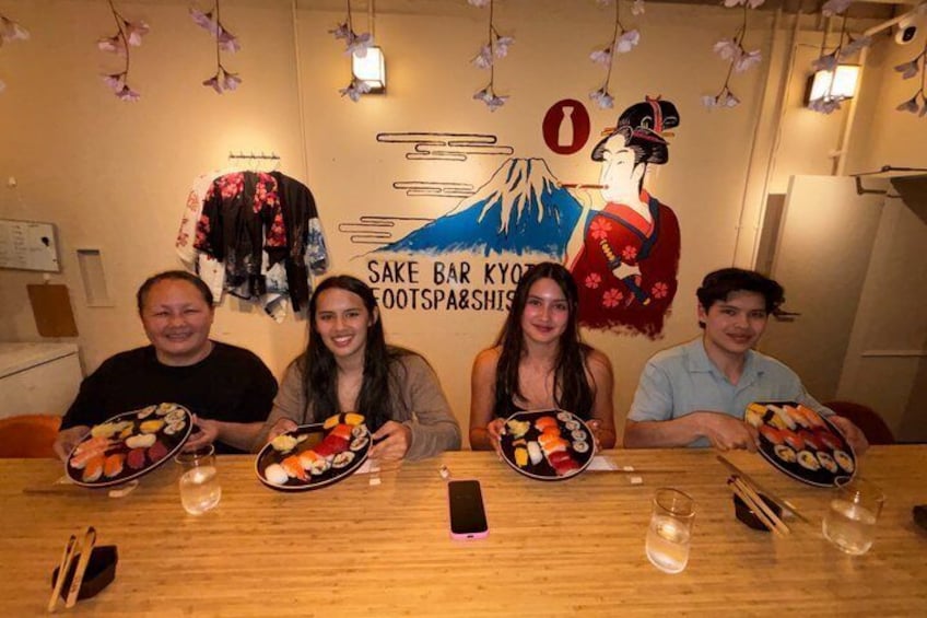 A group of people at a table with plates of sushi, a mural with Japanese art, and a sign that reads 'SAKE BAR KYOR FOOTSPARSHIS.' 4