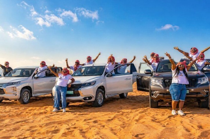 Multiple people in white shirts and red headscarves posing with cars in a desert. 1