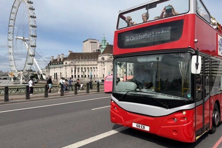 A red double-decker bus with 'Christopher' written on the front, driving on a bridge with a Ferris wheel in the background. 1