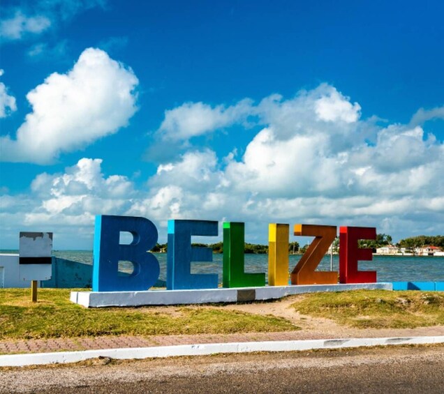 The word 'BELIZE' in colorful letters on a grassy area with a body of water and a cloudy sky in the background. 3