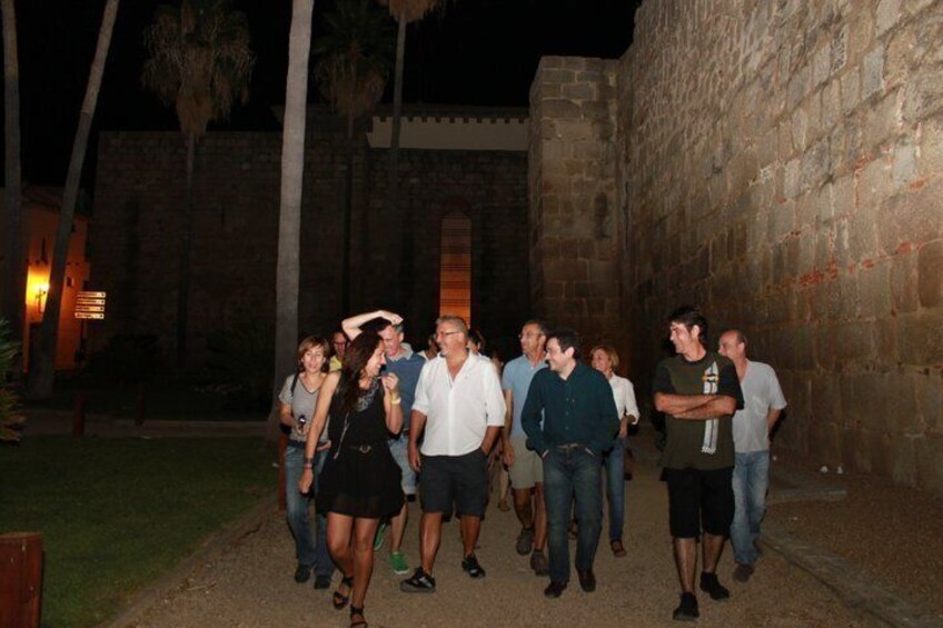 A group of people walking along a path at night in an outdoor setting with stone walls and palm trees. 5