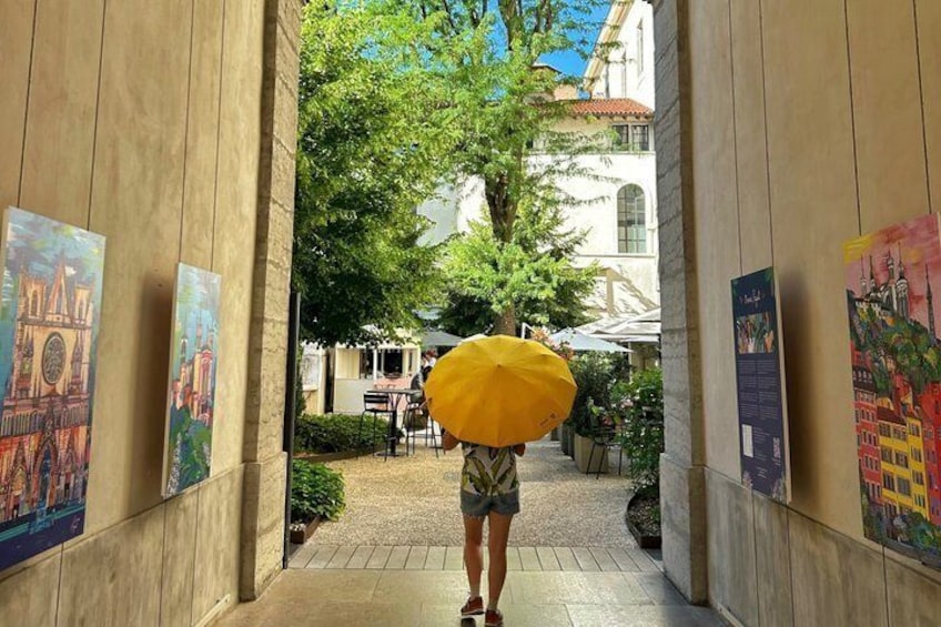 A person walking down a corridor with paintings on the walls, a yellow umbrella, and a building in the background. 1