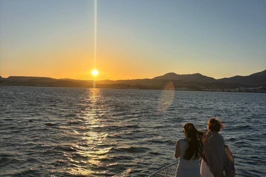 Two people standing on a boat, facing a sunset over a body of water with mountains in the background. 4