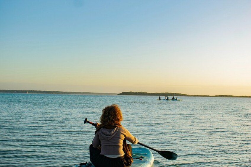 A person kayaking on calm water with a distant shoreline and a small boat in the background. 3