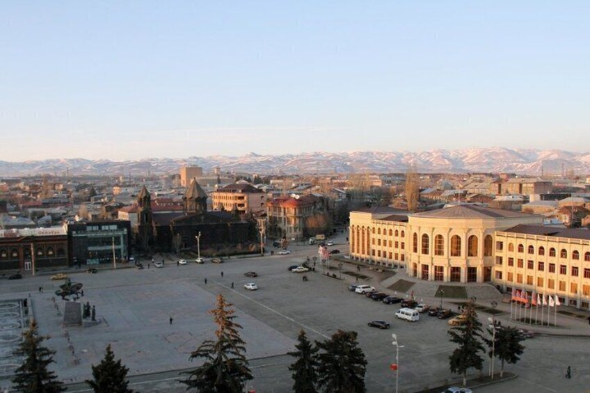 A cityscape with a prominent building with arched windows, a large open square, and snow-capped mountains in the background. 4