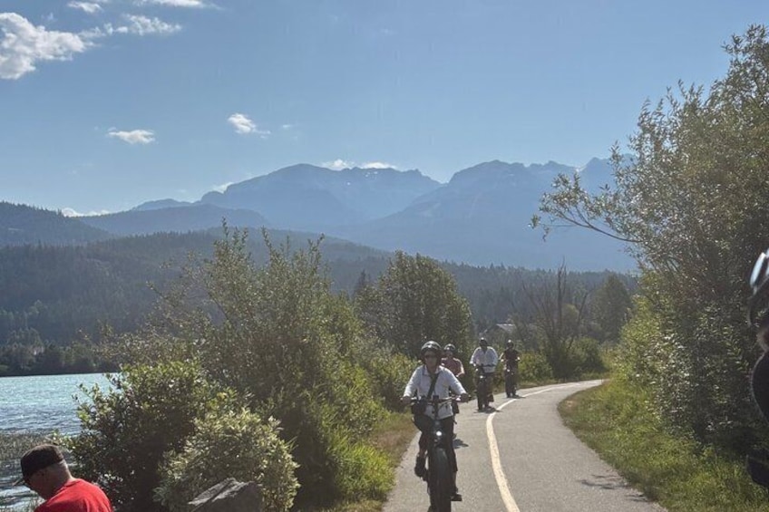 A group of cyclists on a paved path with a lake and mountains in the background. 2