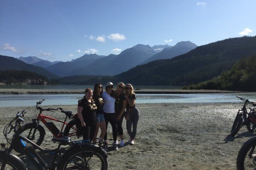A group of people standing by a lake with mountains in the background and bicycles nearby. 3