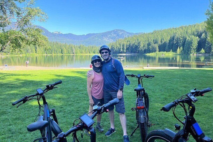 Two people standing with bicycles in a grassy area near a lake with mountains in the background. 1