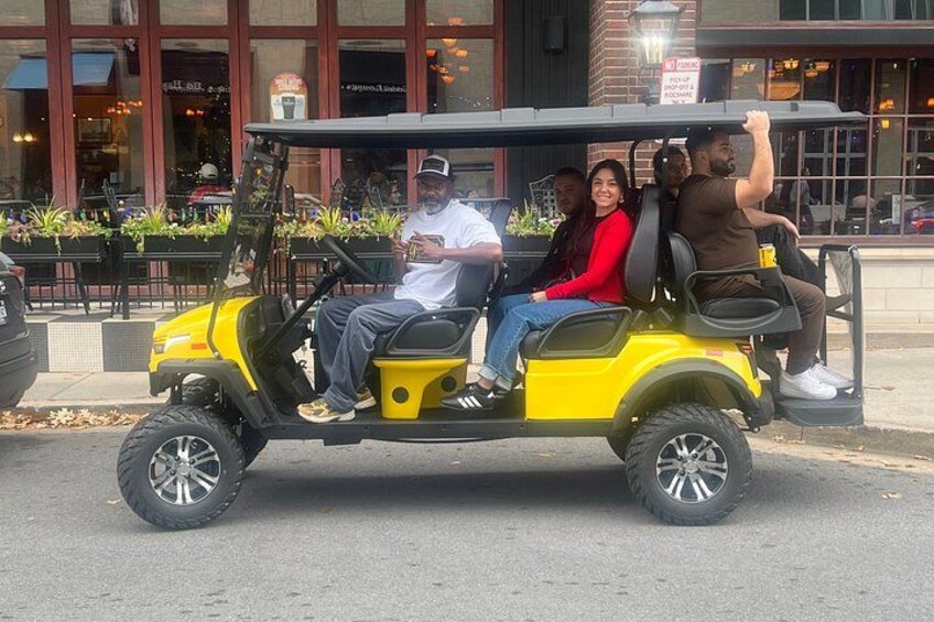 A yellow golf cart with four passengers on a street. 1