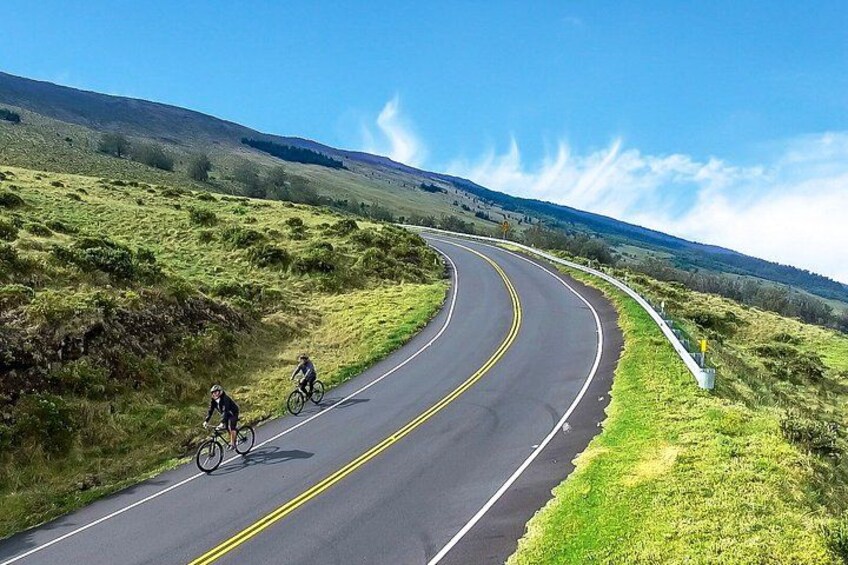 A curving road with cyclists, surrounded by grassy hills and a clear sky. 1