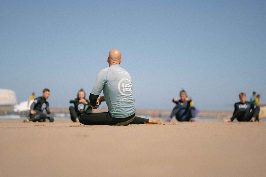 A man in a light blue shirt sitting on the beach with a logo on the back, surrounded by people in wetsuits. 1