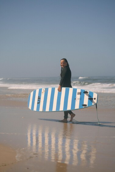 A person holding a blue and white striped surfboard on a beach with waves in the background. 2