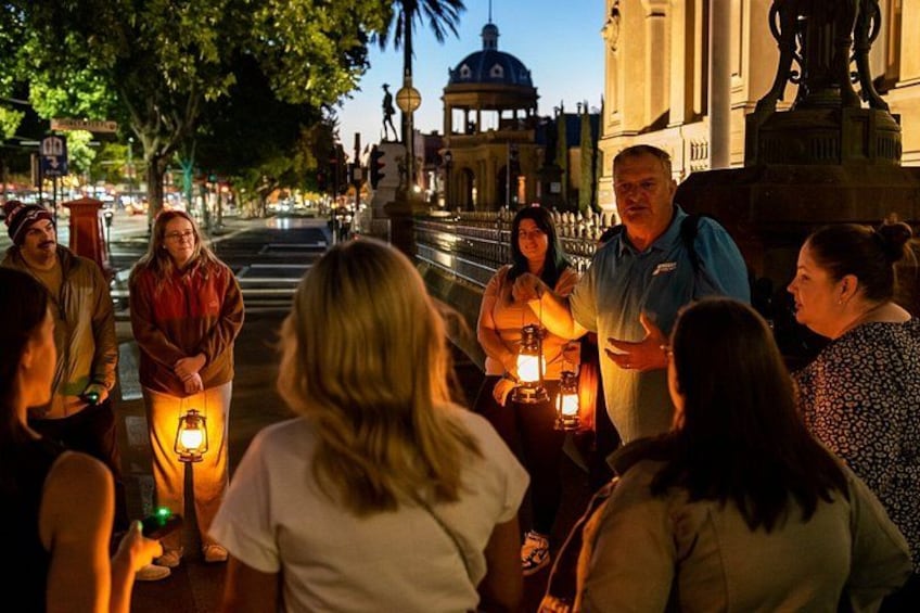 A group of people gathered around a street lamp at night, with buildings and a dome structure in the background. 1