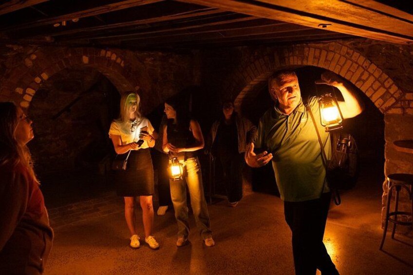 A group of people in a dimly lit underground setting with brick arches, a person holding a lantern, and a stool visible. 5