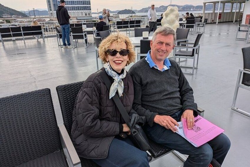 To personer sidder på en terrasse med stole og et bord, med udsigt til byens skyline i baggrunden. 3