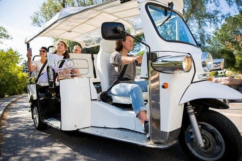 A white golf cart with passengers, parked on a paved path with greenery in the background. 1