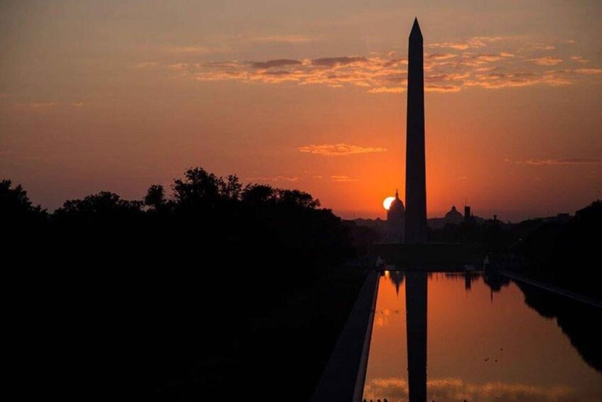 A sunset with a tall monument reflecting on water, surrounded by trees and a city skyline. 1