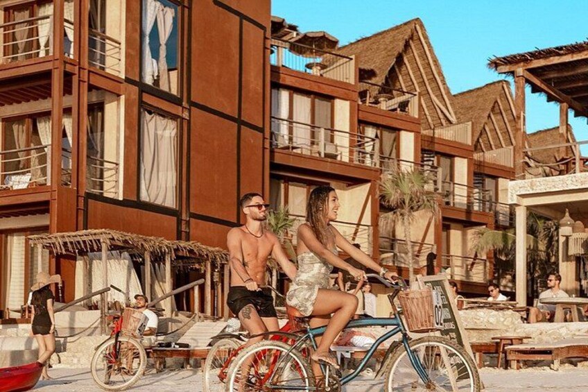 A couple on a bicycle in a resort area with wooden buildings and a beach. 1