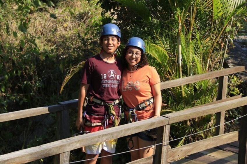 Two people wearing helmets and harnesses on a wooden bridge with greenery in the background. 5